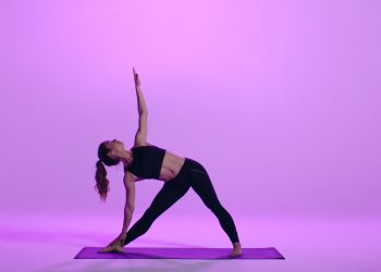 Uma pessoa pratica ioga em um estúdio bem iluminado e roxo. Eles estão realizando a postura do triângulo estendido, esticando um braço em direção ao teto e o outro em direção ao chão, enquanto mantêm uma postura ampla em um tapete de ioga, como parte de seu curso de ioga. | YogaLounge