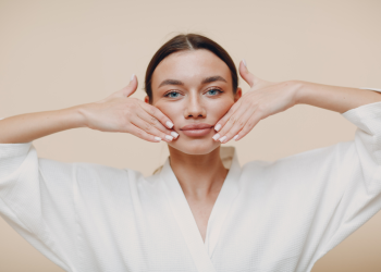 Uma pessoa com longos cabelos escuros e uma túnica branca está realizando um exercício de Yoga Facial com as mãos colocadas em cada lado do rosto. O fundo é de cor bege claro e liso. A pessoa tem uma expressão neutra e parece focada no exercício. | YogaLounge