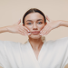 Uma pessoa com longos cabelos escuros e uma túnica branca está realizando um exercício de Yoga Facial com as mãos colocadas em cada lado do rosto. O fundo é de cor bege claro e liso. A pessoa tem uma expressão neutra e parece focada no exercício. | YogaLounge