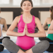 Três mulheres grávidas estão sentadas em posição de lótus em tapetes de Yoga, com os olhos fechados e as mãos em posição de oração. Eles têm roupas de ginástica em cores diferentes: um com regata branca, outro com regata rosa e o terceiro com regata verde. | YogaLounge