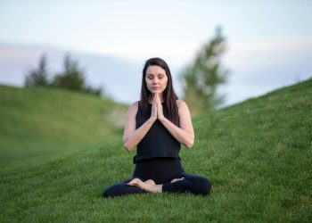 Uma pessoa sentada de pernas cruzadas na relva verde, olhos fechados e mãos em posição de oração junto ao peito, celebrando o Dia Internacional do Yoga. Ela veste um top preto sem mangas e calça preta, com uma paisagem de árvores e céu azul ao fundo. | YogaLounge