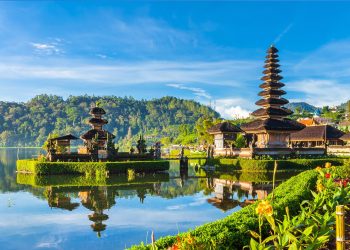 Uma cena pitoresca do Templo Ulun Danu Bratan em Bali, Indonésia, situado perto de um lago sereno com reflexos claros do templo e das árvores verdes exuberantes ao redor. O céu é de um azul vibrante com nuvens dispersas e flores coloridas em primeiro plano - um cenário perfeito para um retiro do Curso Yoga Bali. | YogaLounge