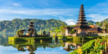 Uma cena pitoresca do Templo Ulun Danu Bratan em Bali, Indonésia, situado perto de um lago sereno com reflexos claros do templo e das árvores verdes exuberantes ao redor. O céu é de um azul vibrante com nuvens dispersas e flores coloridas em primeiro plano - um cenário perfeito para um retiro do Curso Yoga Bali. | YogaLounge