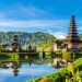 Uma cena pitoresca do Templo Ulun Danu Bratan em Bali, Indonésia, situado perto de um lago sereno com reflexos claros do templo e das árvores verdes exuberantes ao redor. O céu é de um azul vibrante com nuvens dispersas e flores coloridas em primeiro plano - um cenário perfeito para um retiro do Curso Yoga Bali. | YogaLounge