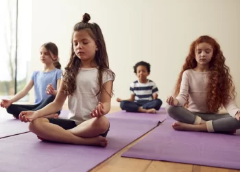 Quatro crianças estão sentadas de pernas cruzadas em tapetes de ioga, em pose meditativa, com os olhos fechados. Eles estão dentro de uma sala iluminada e com parede branca, participando de um “Curso de Yoga para Crianças”. As crianças parecem calmas e focadas. | YogaLounge