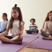 Quatro crianças estão sentadas de pernas cruzadas em tapetes de ioga, em pose meditativa, com os olhos fechados. Eles estão dentro de uma sala iluminada e com parede branca, participando de um “Curso de Yoga para Crianças”. As crianças parecem calmas e focadas. | YogaLounge