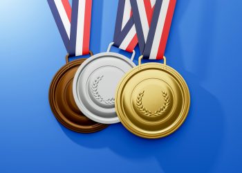 Três medalhas penduradas sobre um fundo azul: uma medalha de bronze à esquerda, uma medalha de prata no meio e uma medalha de ouro à direita. Cada medalha dos Jogos Olímpicos apresenta um desenho de coroa de louros e é presa a uma fita listrada de vermelho, branco e azul. | YogaLounge