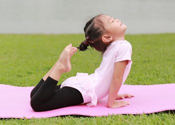 Uma jovem praticando ioga ao ar livre em um tapete rosa, realizando a postura do arco. Ela está vestindo uma camisa branca e legging preta, com as mãos segurando os pés atrás das costas. O fundo é uma área gramada com um corpo d'água visível ao longe, parte de seu curso de yoga infantil. | YogaLounge