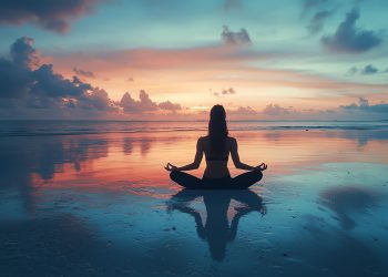 Uma silhueta de uma pessoa sentada de pernas cruzadas em uma pose meditativa em uma praia de areia molhada ao pôr do sol. O céu está vibrante com tons de laranja, rosa e azul, e a água calma cria um reflexo espelhado da cena, aumentando a atmosfera tranquila de paz. | YogaLounge