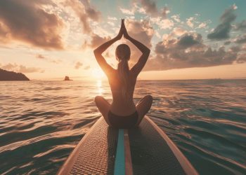 Uma pessoa senta-se de pernas cruzadas em uma prancha de remo, realizando uma pose de yoga com as mãos levantadas acima da cabeça ao pôr do sol. O sol cria um brilho quente e dourado na água, e as nuvens se espalham pelo horizonte na mistura definitiva de surfe e serenidade de yoga. | YogaLounge