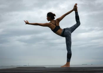 Uma pessoa executa a pose de yoga do dançarino em uma plataforma de madeira contra um céu nublado, criando uma experiência de equilíbrio e graça. Elas ficam em uma perna, estendendo a outra perna e um braço para trás enquanto alcançam a frente com o outro braço, vestidas com um sutiã esportivo preto e leggings estampadas. | YogaLounge
