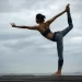 Uma pessoa executa a pose de yoga do dançarino em uma plataforma de madeira contra um céu nublado, criando uma experiência de equilíbrio e graça. Elas ficam em uma perna, estendendo a outra perna e um braço para trás enquanto alcançam a frente com o outro braço, vestidas com um sutiã esportivo preto e leggings estampadas. | YogaLounge