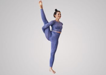 Uma pessoa em uma roupa de yoga roxa faz uma pose de split em pé contra um fundo claro simples. Ela fica em pé sobre uma perna com a outra perna estendida para cima, segurando a perna levantada com uma mão. Ela tem uma expressão serena e seu cabelo está preso, incorporando 20 anos de experiência e confiança como referência em formação de professores de yoga. | YogaLounge