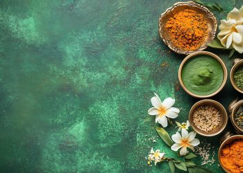 Várias tigelas de especiarias coloridas são dispostas em uma superfície verde texturizada, incorporando a essência do Ayurveda. As tigelas contêm especiarias laranja, verde e marrom. Flores brancas são colocadas entre as tigelas, adicionando um toque fresco à composição harmoniosa. | YogaLounge