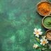 Várias tigelas de especiarias coloridas são dispostas em uma superfície verde texturizada, incorporando a essência do Ayurveda. As tigelas contêm especiarias laranja, verde e marrom. Flores brancas são colocadas entre as tigelas, adicionando um toque fresco à composição harmoniosa. | YogaLounge