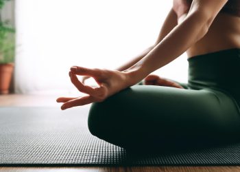 Uma pessoa senta-se de pernas cruzadas sobre um tapete de yoga em uma pose meditativa, com uma mão apoiada no joelho, formando um mudra. O fundo tem iluminação suave e algumas plantas de interior, evocando o espírito tranquilo da História do Yoga. A atmosfera é calma e relaxante. | YogaLounge