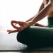 Uma pessoa senta-se de pernas cruzadas sobre um tapete de yoga em uma pose meditativa, com uma mão apoiada no joelho, formando um mudra. O fundo tem iluminação suave e algumas plantas de interior, evocando o espírito tranquilo da História do Yoga. A atmosfera é calma e relaxante. | YogaLounge