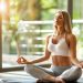 Uma mulher senta-se de pernas cruzadas em um tapete de ioga em uma sala ensolarada, praticando Terapia de Atenção Plena com os olhos fechados. Ela usa um sutiã esportivo branco e leggings cinza. Plantas em vasos e grandes janelas estão ao fundo, realçando a atmosfera serena. | YogaLounge