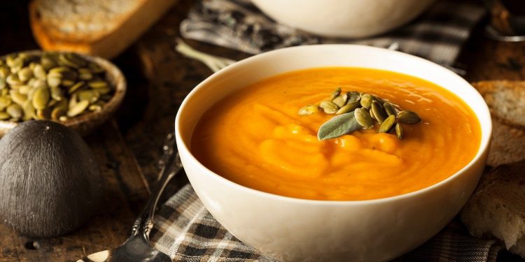 Uma tigela de sopa cremosa de abóbora guarnecida com sementes de abóbora e uma folha de sálvia. Uma colher e uma fatia de pão estão ao lado. Outra tigela de sopa é parcialmente visível ao fundo em uma mesa de madeira rústica. | YogaLounge
