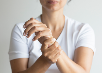 Uma pessoa de camisa branca é mostrada segurando o pulso esquerdo com a mão direita, sugerindo desconforto potencialmente ligado à osteoporose. O fundo permanece simples e discreto. | YogaLounge