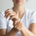 Uma pessoa de camisa branca é mostrada segurando o pulso esquerdo com a mão direita, sugerindo desconforto potencialmente ligado à osteoporose. O fundo permanece simples e discreto. | YogaLounge