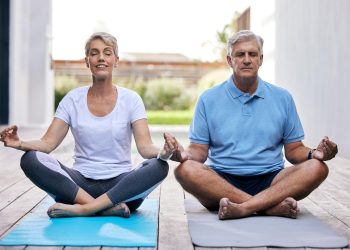 Um homem e uma mulher sentam-se de pernas cruzadas em tapetes de ioga, meditando ao ar livre em um deck de madeira durante um retiro de yoga. Eles parecem relaxados e focados, com árvores ao fundo. Ambos estão vestindo roupas confortáveis: a mulher com uma blusa branca e o homem com uma camisa polo azul.
 | YogaLounge