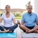 Um homem e uma mulher sentam-se de pernas cruzadas em tapetes de ioga, meditando ao ar livre em um deck de madeira durante um retiro de yoga. Eles parecem relaxados e focados, com árvores ao fundo. Ambos estão vestindo roupas confortáveis: a mulher com uma blusa branca e o homem com uma camisa polo azul.
| YogaLounge