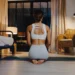Uma mulher está ajoelhada em um tapete de yoga em uma sala mal iluminada, incorporando a prática de yoga para um sono mais profundo. Ela usa um sutiã esportivo branco e shorts. O espaço tranquilo apresenta uma cama, cadeira de vime, luminária e janela com cortinas. | YogaLounge