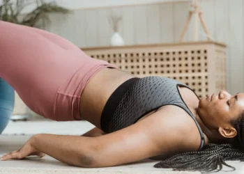 Uma pessoa demonstrando um exercício de ponte em um tapete dentro de casa, usando um sutiã esportivo cinza e leggings rosas, explora como emagrecer com yoga. O quarto apresenta decoração suave e neutra com um armário de vime e plantas exuberantes ao fundo. | YogaLounge