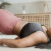 Uma pessoa demonstrando um exercício de ponte em um tapete dentro de casa, usando um sutiã esportivo cinza e leggings rosas, explora como emagrecer com yoga. O quarto apresenta decoração suave e neutra com um armário de vime e plantas exuberantes ao fundo. | YogaLounge