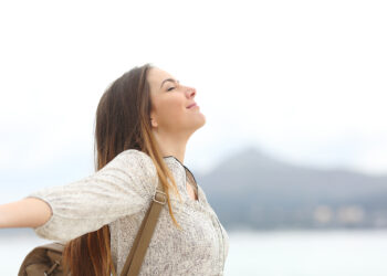 Uma mulher está ao ar livre com os braços estendidos, olhos fechados e uma expressão serena, como se estivesse liberando toda a ansiedade. Ela está usando um suéter leve e carregando uma mochila contra o fundo desfocado de montanhas e um céu nublado. | YogaLounge