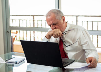 Homem idoso de camisa branca e gravata vermelha sentado à mesa com um laptop, segurando óculos e cobrindo a boca com a mão. O olhar de cansaço extremo é evidente, pois ele parece pensativo, olhando para papéis, com uma janela ao fundo mostrando uma grade de sacada. | YogaLounge