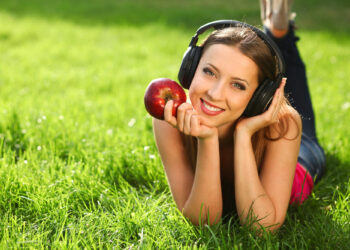 Uma mulher está deitada na relva, sorrindo e usando fones de ouvido. Abraçando um estilo de vida saudável, ela segura uma maçã vermelha em uma mão enquanto apoia o queixo na outra. Com cabelos longos e roupas casuais, a cena brilhante e ensolarada transmite uma atmosfera relaxada. | YogaLounge