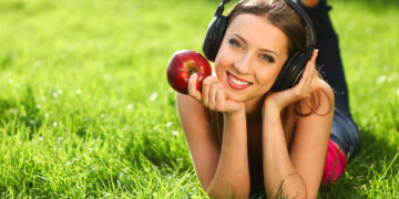 Uma mulher está deitada na relva, sorrindo e usando fones de ouvido. Abraçando um estilo de vida saudável, ela segura uma maçã vermelha em uma mão enquanto apoia o queixo na outra. Com cabelos longos e roupas casuais, a cena brilhante e ensolarada transmite uma atmosfera relaxada. | YogaLounge