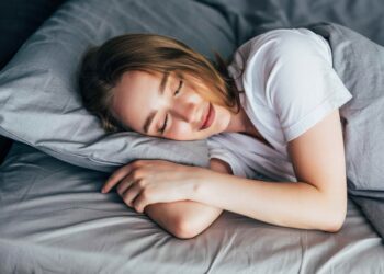 Uma pessoa com cabelos longos dorme pacificamente em uma cama cinza, vestindo uma camiseta branca. Ela está deitada de lado com a cabeça apoiada em um travesseiro e os braços dobrados sob ele. A iluminação suave e a expressão relaxada sugerem um descanso confortável. | YogaLounge