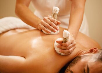 Uma pessoa se entrega a uma massagem de luxo com compressas de ervas nas costas. Mãos habilidosas pressionam bolsas de pano cheias de ervas calmantes na pele, deitada de bruços em uma mesa de massagem em um ambiente de bem-estar quente e sereno. | YogaLounge