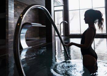 Silhueta de uma mulher em pé, com água até a cintura, em uma piscina coberta, perto de um corrimão curvo. Ela está usando um maiô, e a cena exala bem-estar deluxe com sua iluminação suave de luz natural que flui através de grandes janelas ao fundo. | YogaLounge