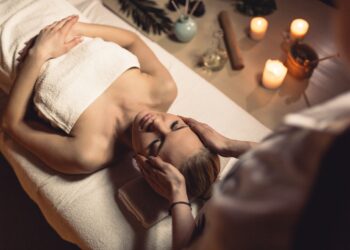 Uma pessoa desfruta de uma massagem deluxe na cabeça em uma mesa aconchegante, enrolada em uma toalha. A sala suavemente iluminada, adornada com velas e itens aromáticos, cria um ambiente de calma e bem-estar. | YogaLounge
