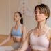 Duas mulheres meditam em uma sala tranquila, sentadas de pernas cruzadas em tapetes de ioga. Vestidas com roupas de ginástica confortáveis, elas fecham os olhos e se concentram internamente. Um gráfico de chakras enfeita a parede do fundo, aumentando a atmosfera serena que oferece consolo durante as transições da menopausa. | YogaLounge