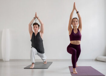 Um homem e uma mulher praticam yoga em tapetes em uma sala iluminada. Ambos estão em uma pose de yoga em pé com uma perna dobrada e as mãos levantadas acima de suas cabeças. A mulher usa uma roupa marrom, e o homem usa uma blusa preta e calças brancas. | YogaLounge