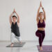Um homem e uma mulher praticam yoga em tapetes em uma sala iluminada. Ambos estão em uma pose de yoga em pé com uma perna dobrada e as mãos levantadas acima de suas cabeças. A mulher usa uma roupa marrom, e o homem usa uma blusa preta e calças brancas. | YogaLounge