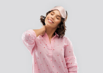 Mulher sorrindo com os olhos fechados, usando uma máscara de dormir rosa no topo da cabeça e uma camisa de pijama rosa com pequenos corações vermelhos. Relaxando como se tivesse acabado de comparar os efeitos de Victan Para Dormir vs Yoga, ela toca a parte de trás do pescoço com uma mão, contra um fundo cinza simples. | YogaLounge