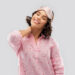 Mulher sorrindo com os olhos fechados, usando uma máscara de dormir rosa no topo da cabeça e uma camisa de pijama rosa com pequenos corações vermelhos. Relaxando como se tivesse acabado de comparar os efeitos de Victan Para Dormir vs Yoga, ela toca a parte de trás do pescoço com uma mão, contra um fundo cinza simples. | YogaLounge