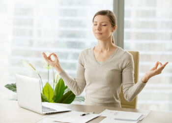 Uma pessoa está sentada em uma mesa com os olhos fechados, praticando meditação com as mãos em uma pose mudra. Um laptop e papéis estão sobre a mesa. Plantas verdes são visíveis no fundo com uma vista de janela desfocada. | YogaLounge