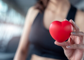 Em um ambiente de academia, uma pessoa usando um sutiã esportivo preto segura um pequeno objeto vermelho em forma de coração em foco, simbolizando saúde cardiovascular. O fundo desfocado enfatiza ainda mais o coração. | YogaLounge