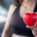Em um ambiente de academia, uma pessoa usando um sutiã esportivo preto segura um pequeno objeto vermelho em forma de coração em foco, simbolizando saúde cardiovascular. O fundo desfocado enfatiza ainda mais o coração. | YogaLounge