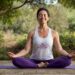 Uma pessoa está sentada de pernas cruzadas em um tapete de yoga roxo ao ar livre, com os olhos fechados e uma expressão pacífica. Ela está vestindo uma regata branca e leggings roxas. Há vegetação exuberante e árvores ao fundo. | YogaLounge