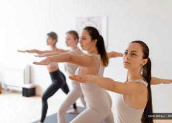 Quatro pessoas estão praticando yoga em ambientes fechados, de pé em uma linha reta e realizando a pose do Guerreiro II com os braços estendidos. Elas vestem roupas esportivas justas e de cor clara. A sala é iluminada e decorada de forma minimalista, lembrando um espaço sereno para aulas de yoga. | YogaLounge