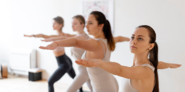 Quatro pessoas estão praticando yoga em ambientes fechados, de pé em uma linha reta e realizando a pose do Guerreiro II com os braços estendidos. Elas vestem roupas esportivas justas e de cor clara. A sala é iluminada e decorada de forma minimalista, lembrando um espaço sereno para aulas de yoga. | YogaLounge