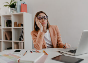 Uma mulher usando óculos e um blazer rosa está sentada em sua mesa, uma mão no rosto, parecendo entediada ou cansada. Em meio a cadernos espalhados e lápis de cor, ela contempla como aumentar a concentração no trabalho através do yoga enquanto prateleiras se avultam ao fundo. | YogaLounge
